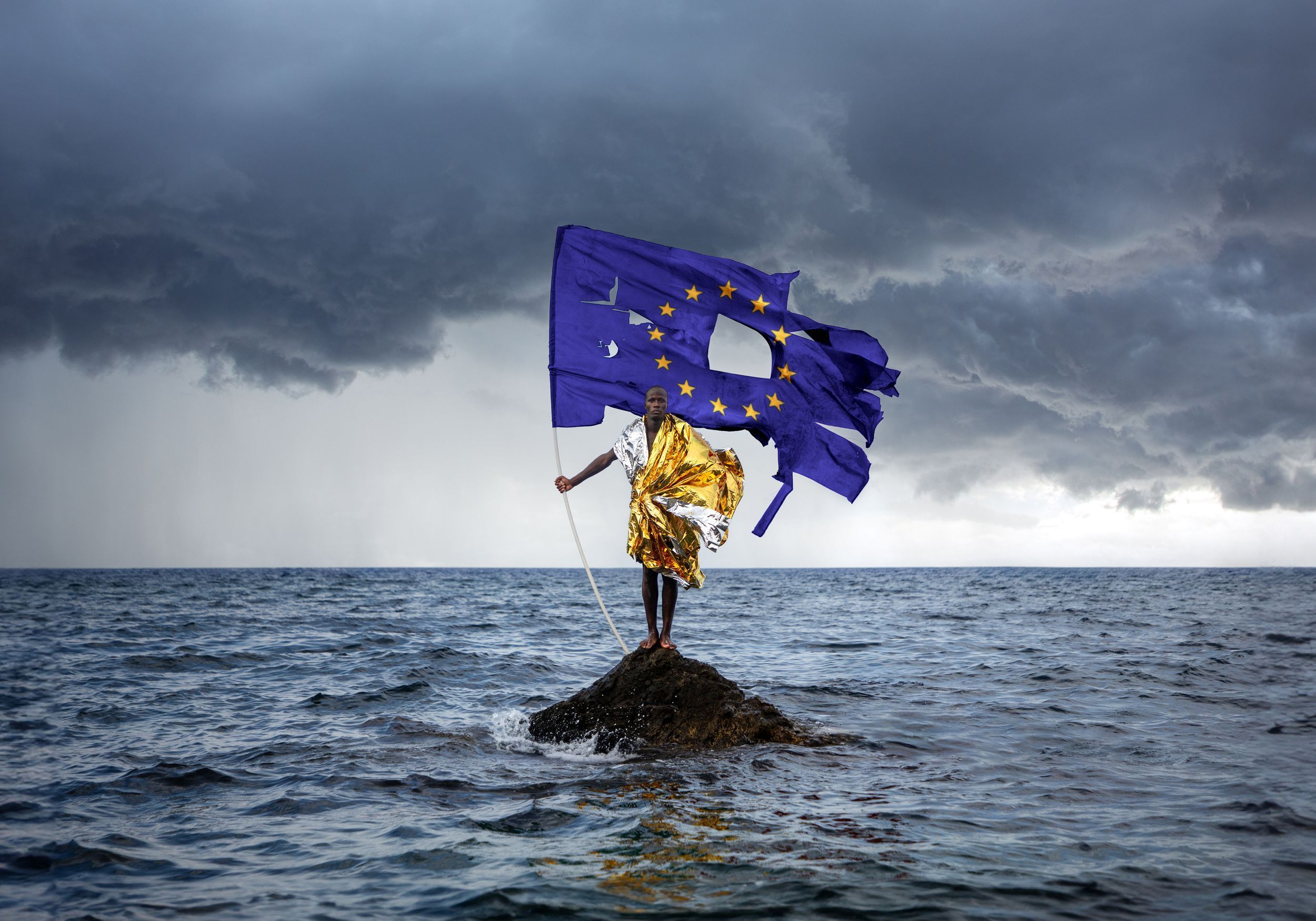 Un migrante su uno scoglio in mezzo al mare con una bandiera europea tutta strappata