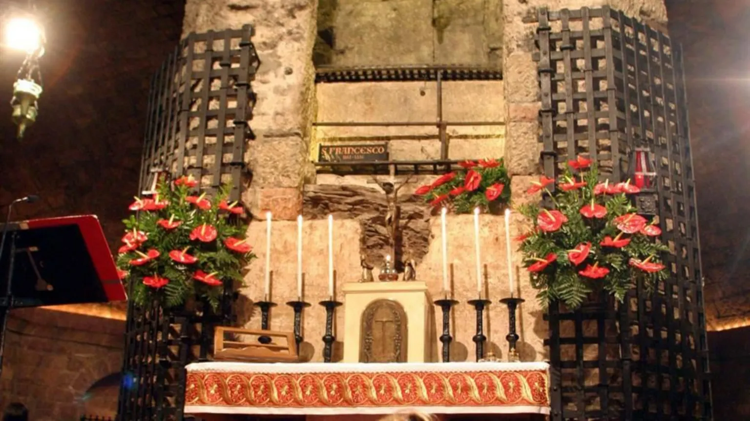 Altare in una cripta di pietra, illuminato in modo soffuso. Davanti ci sono sei candele bianche, un piccolo crocifisso e un rivestimento rosso ricamato. Ai lati, due grandi griglie metalliche con composizioni di fiori rossi. Al centro, in alto, una lapide