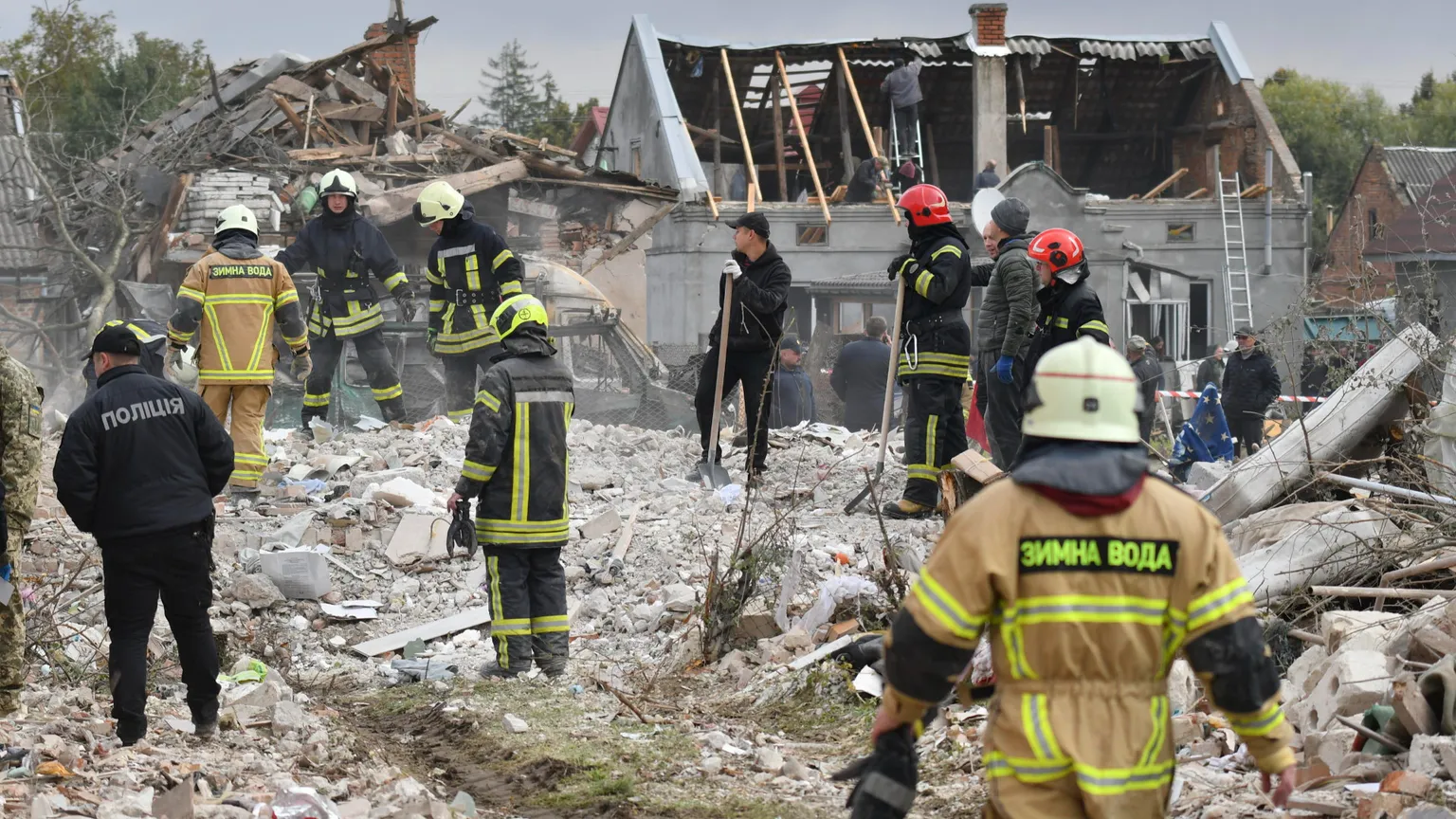 Uomini con tute da soccorritori, casco, guanti e pale in piedi su una distesa di detriti. Sullo sfondo un edificio con tetto a capanna scoperchiato