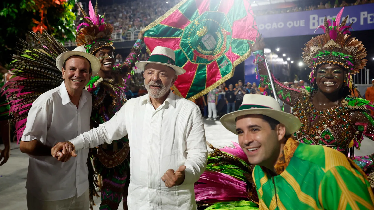 Lula vestito di bianco, con un panama in testa, in mezzo a due uomini col panama: quello a destra avvolto nei colori giallo e verde del Brasile. Sullo sfondo, ballerine in maschera e una bandiera