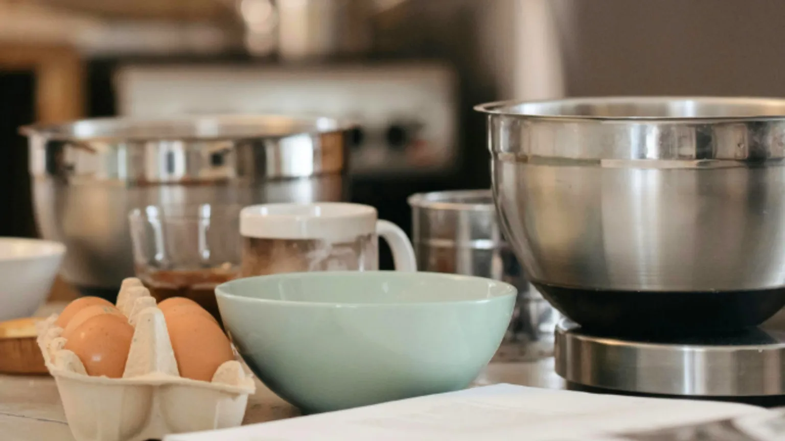 Preparazione di un nuovo piatto. Si vedono, su un tavolo, quattro contenitori, uno di ceramica e tre di acciaio, e diverse uova. In primo piano, sfocato, un libro di ricette