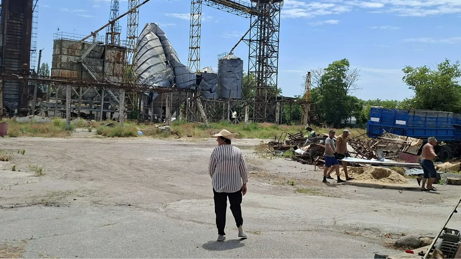 Un uomo è in piedi al centro di una strada. Sullo sfondo un silos semi distrutto. Altri due uomini appaiono a destra nella scena