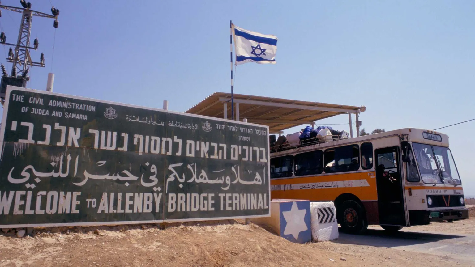 Un cartellone verde, sulla sabbia. Accanto, un autobus colmo di persone. La bandiera israeliana sventola in alto a destra.