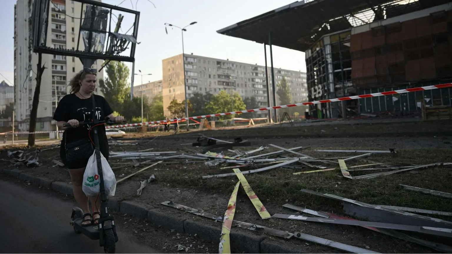 Una ragazza su un monopattino; sullo sfondo, macerie dovute a un bombardamento.