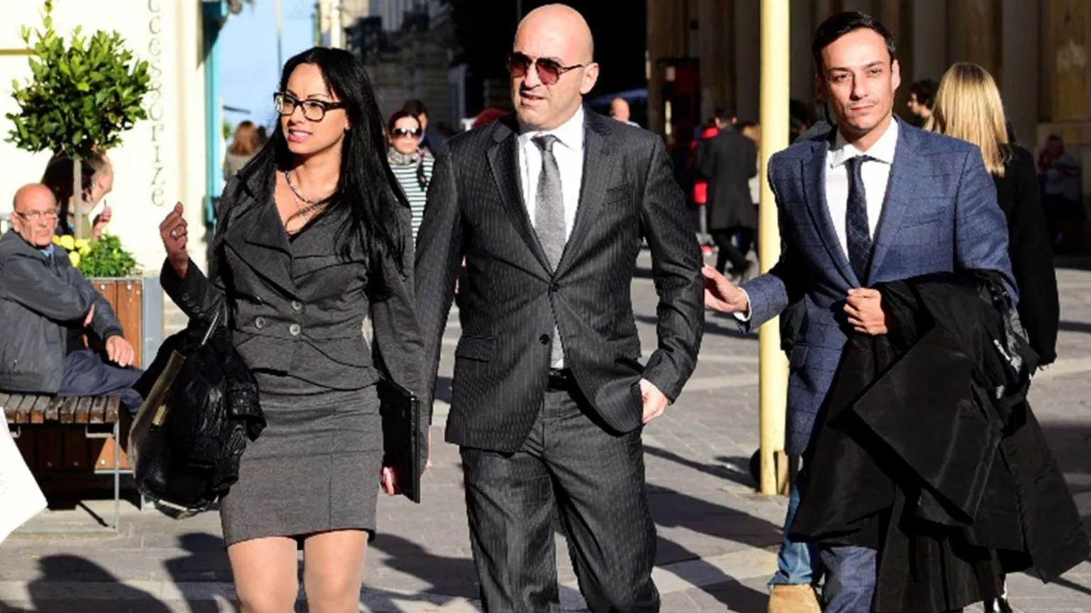 Un uomo di mezza età in completo grigio perla tra una giovane donna in tailleur grigio e un giovane in completo blu