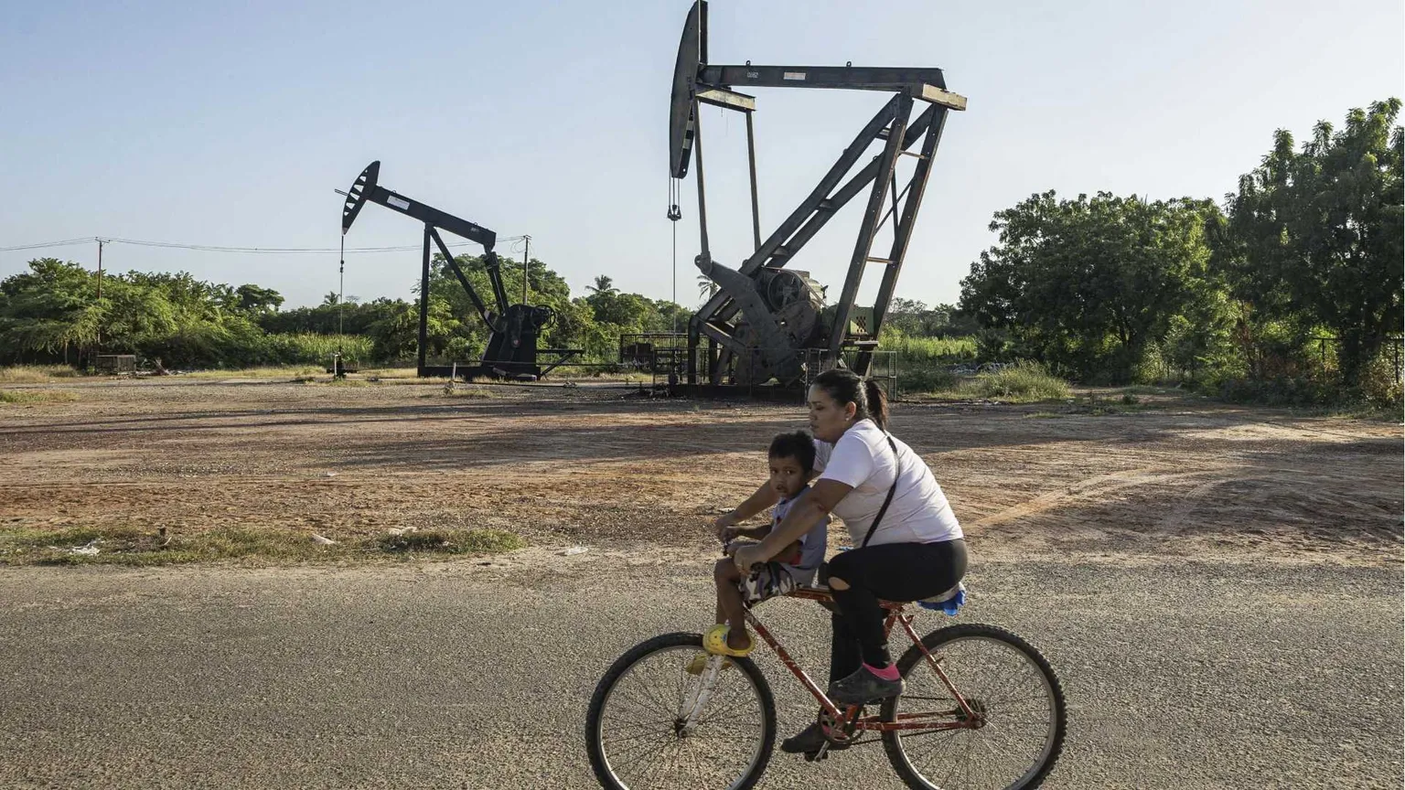 Sullo sfondo due trivelle gigantesche, in primo piano una donna in bicicletta con un bambino sui 3 o 4 anni sulla canna della bici