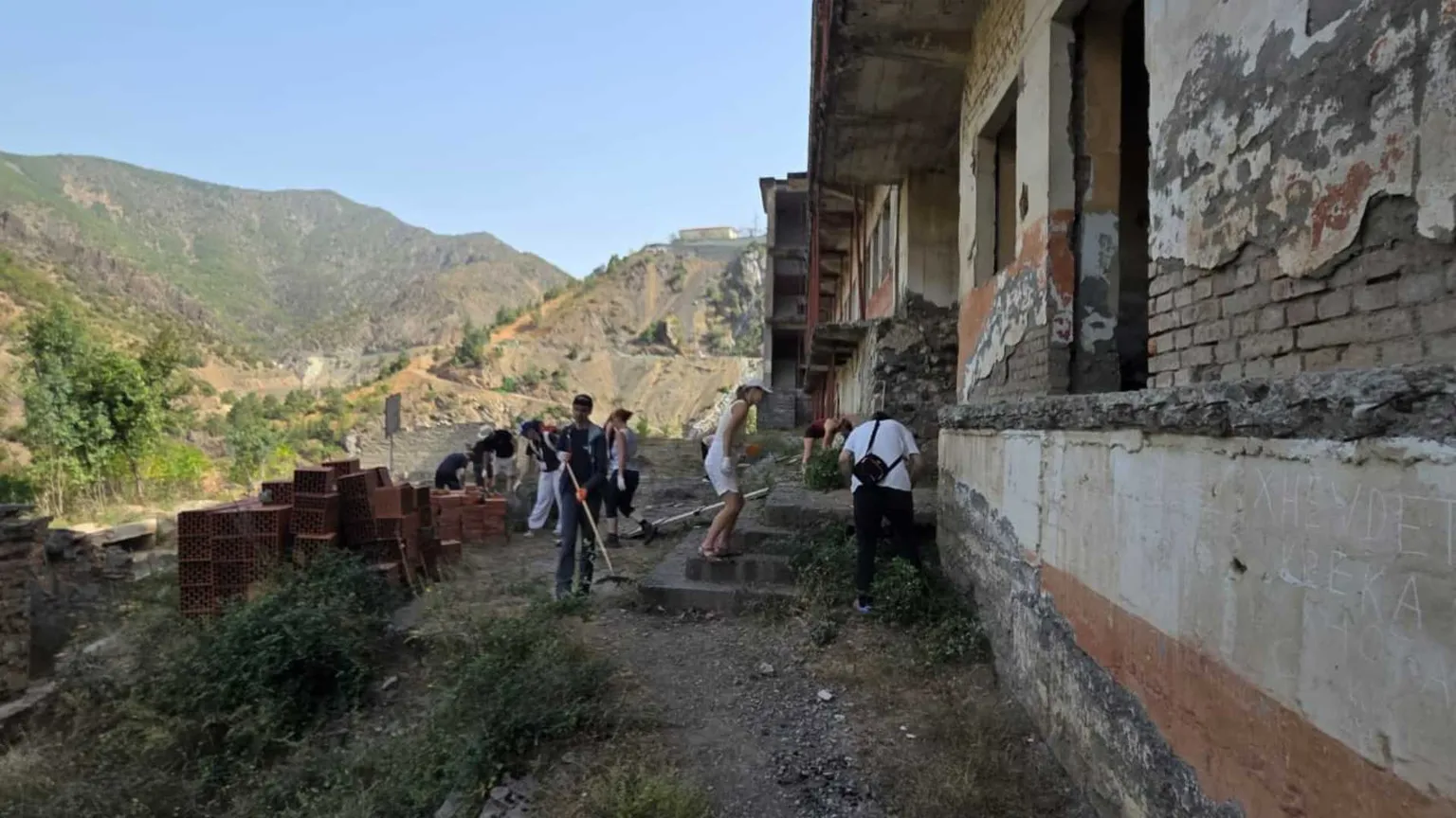 Dei ragazzi muniti di rastrelli e falcette strappano le erbacce di fronte a una prigione abbandonata. Davanti all'edificio giacciono dei mattoni nuovi inutilizzati. Sullo sfondo si vedono le montagne