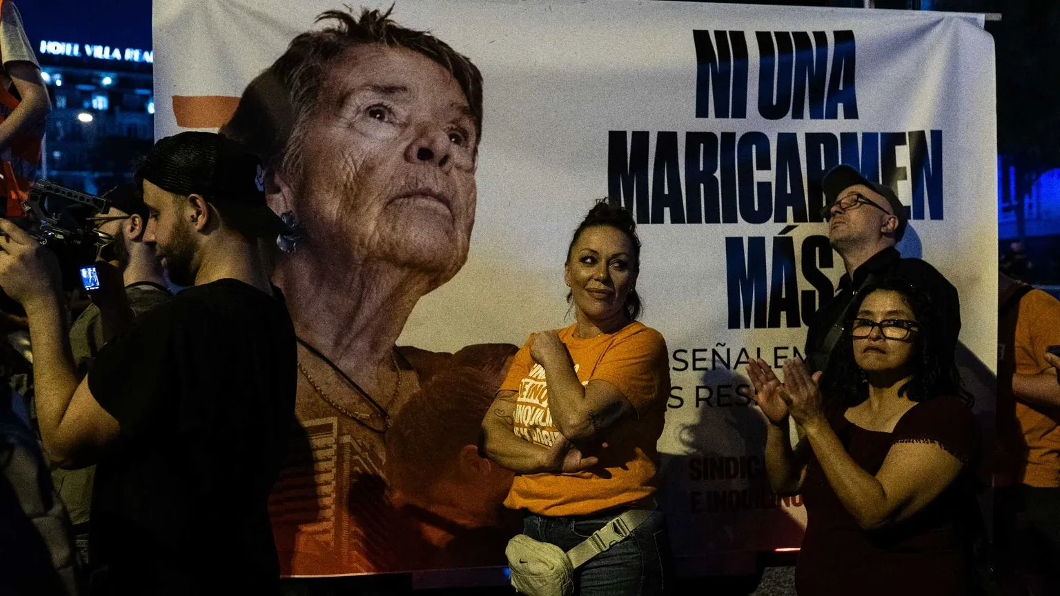 Il volto di una signora anziana su un cartellone con una scritta. Davanti al cartellone ci sono dei manifestanti