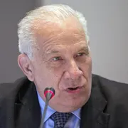 Primo piano di un uomo anziano dai capelli bianchi, in giacca e cravatta scura. È seduto a un tavolo e parla davanti a un microfono con una piccola luce rossa accesa. Lo sfondo è neutro; il suo volto è leggermente di profilo e l’espressione è concentrata.