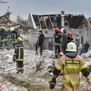 Uomini con tute da soccorritori, casco, guanti e pale in piedi su una distesa di detriti. Sullo sfondo un edificio con tetto a capanna scoperchiato