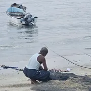 Un uomo in canottiera e a piedi nudi è accucciato sulla battigia davanti a reti da pesca