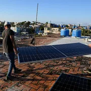 Due uomini sono sul tetto di una casa e trasportano un pannello solare. Dovranno installarlo in cima all'edificio. Sullo sfondo si intravedono altre case, alle cui sommità ci sono alcune cisterne per conservare l'acqua piovana.
