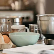 Preparazione di un nuovo piatto. Si vedono, su un tavolo, quattro contenitori, uno di ceramica e tre di acciaio, e diverse uova. In primo piano, sfocato, un libro di ricette