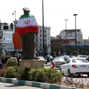 Una rotonda trafficata con, al centro, un'alta statua di un uomo con turbante: sulle spalle la bandiera dell'Iran