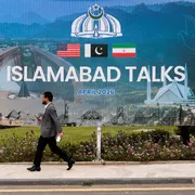 Un uomo passa davanti a un gigantesco cartellone con la scritta "Islamabad Talks" e le bandiere di Usa, Pakistan e Iran sormontate dall'emblema del Pakistan, una corona con mezzaluna