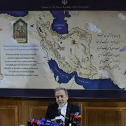 Un uomo in giacca nera e camicia biancia, capelli e barba brizzolati, seduto davanti a una selva di microfoni: alle sue spalle una mappa del Medio Oriente e, ai lati, le bandiere iraniana e indiana