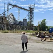Un uomo è in piedi al centro di una strada. Sullo sfondo un silos semi distrutto. Altri due uomini appaiono a destra nella scena