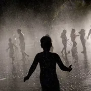 In primo piano la sagoma di un bambino che ha le braccia aperte. Sullo sfondo, sfocate, persone che giocano con vapore d'acqua