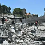 Tre persone camminano tra le macerie di un edificio bombardato. Sullo sfondo, il cielo azzurro