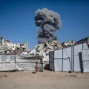 Una colonna di fumo si vede sullo sfondo; davanti, tende degli sfollati. In primo piano, un bambino che corre e scappa dal bombardamento
