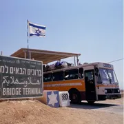Un cartellone verde, sulla sabbia. Accanto, un autobus colmo di persone. La bandiera israeliana sventola in alto a destra.