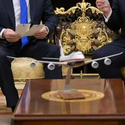 Un modellino di un aereo in primo piano. Sullo sfondo due uomini in abiti eleganti che parlano