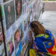 Una giovane donna bionda avvolta in una bandiera gialla e blu con la testa appoggiata contro un muro al quale sono attaccati manifestini con volti di giovani in divisa