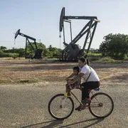 Sullo sfondo due trivelle gigantesche, in primo piano una donna in bicicletta con un bambino sui 3 o 4 anni sulla canna della bici