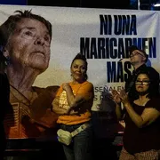 Il volto di una signora anziana su un cartellone con una scritta. Davanti al cartellone ci sono dei manifestanti