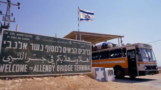 Un cartellone verde, sulla sabbia. Accanto, un autobus colmo di persone. La bandiera israeliana sventola in alto a destra.