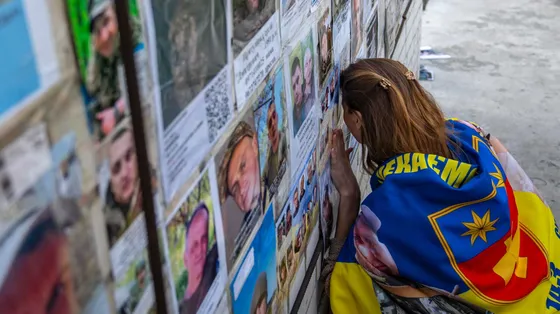 Una giovane donna bionda avvolta in una bandiera gialla e blu con la testa appoggiata contro un muro al quale sono attaccati manifestini con volti di giovani in divisa
