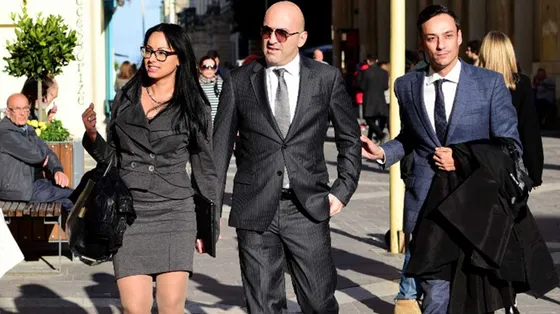 Un uomo di mezza età in completo grigio perla tra una giovane donna in tailleur grigio e un giovane in completo blu