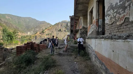 Dei ragazzi muniti di rastrelli e falcette strappano le erbacce di fronte a una prigione abbandonata. Davanti all'edificio giacciono dei mattoni nuovi inutilizzati. Sullo sfondo si vedono le montagne