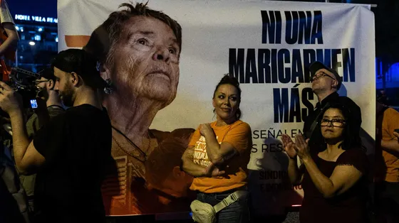 Il volto di una signora anziana su un cartellone con una scritta. Davanti al cartellone ci sono dei manifestanti