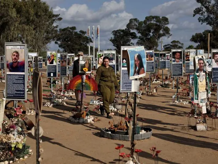 Campo commemorativo all’aperto: su terriccio chiaro sono disposti in file molti pannelli con foto e brevi testi delle vittime, circondati da piccoli memoriali con fiori, candele e oggetti personali