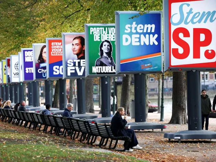 Una persona seduta su una panchina in un parco osserva dei grandi cartelloni elettorali con i simboli e gli slogan dei partiti