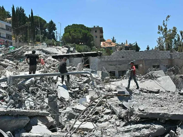 Tre persone camminano tra le macerie di un edificio bombardato. Sullo sfondo, il cielo azzurro