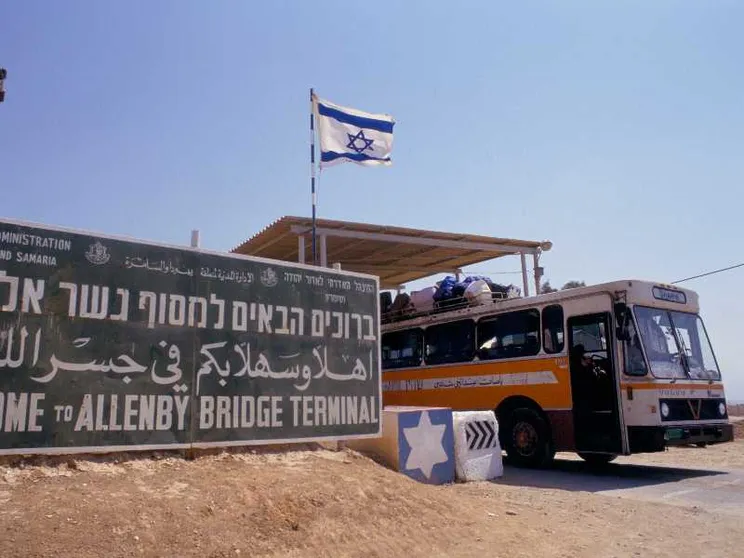 Un cartellone verde, sulla sabbia. Accanto, un autobus colmo di persone. La bandiera israeliana sventola in alto a destra.