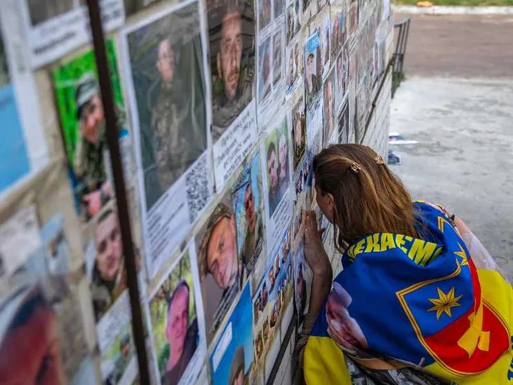 Una giovane donna bionda avvolta in una bandiera gialla e blu con la testa appoggiata contro un muro al quale sono attaccati manifestini con volti di giovani in divisa