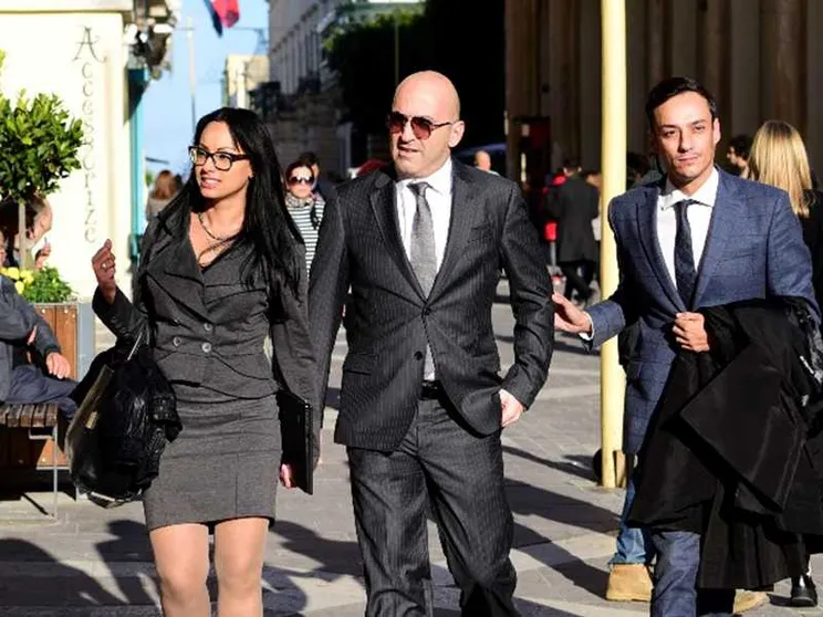 Un uomo di mezza età in completo grigio perla tra una giovane donna in tailleur grigio e un giovane  in completo blu