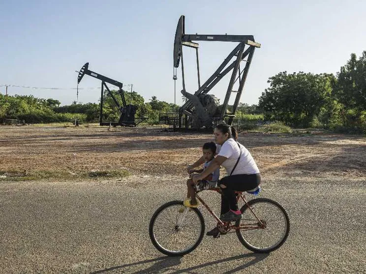 Sullo sfondo due trivelle gigantesche, in primo piano una donna in bicicletta con un bambino sui 3 o 4 anni sulla canna della bici