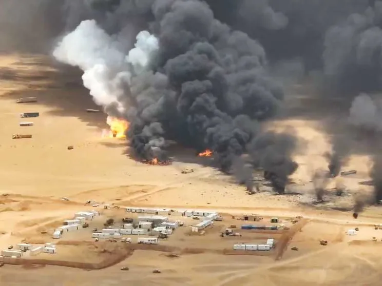 Basse costruzioni bianche in mezzo al deserto di sabbia ocra, sullo sfondo il fumo nerissimo dei bombardamenti