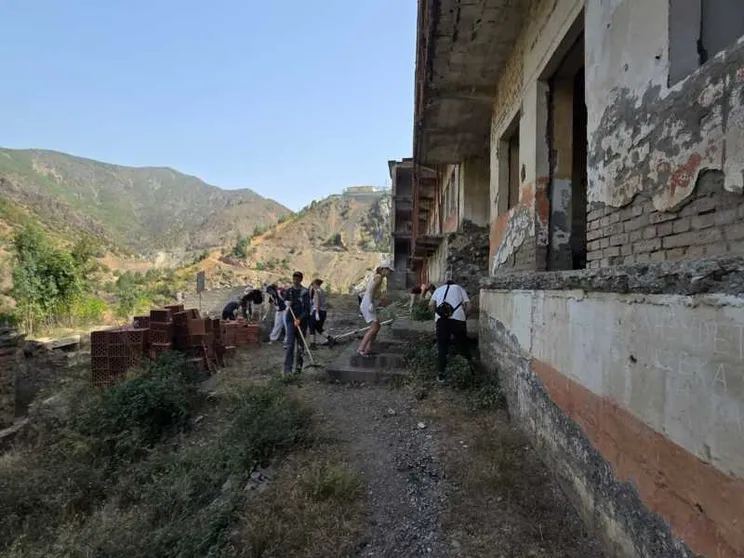 Dei ragazzi muniti di rastrelli e falcette strappano le erbacce di fronte a una prigione abbandonata. Davanti all'edificio giacciono dei mattoni nuovi inutilizzati. Sullo sfondo si vedono le montagne