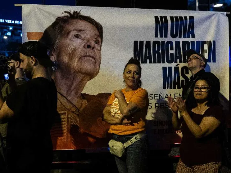 Il volto di una signora anziana su un cartellone con una scritta. Davanti al cartellone ci sono dei manifestanti