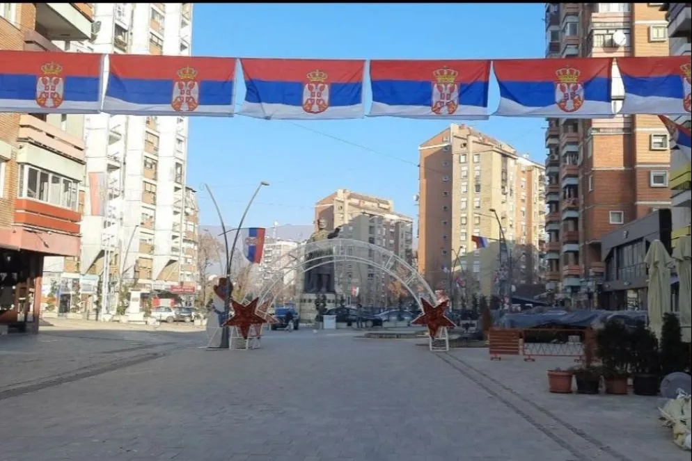 La via principale della parte serba di Kosovska Mitrovica, dove alle bandiere serbe si aggiungono gli addobbi natalizi. Sullo sfondo, la statua di Lazar Hrebeljanović, eroe della battaglia di battaglia di Kosovo Polje, rivolta verso la parte albanese