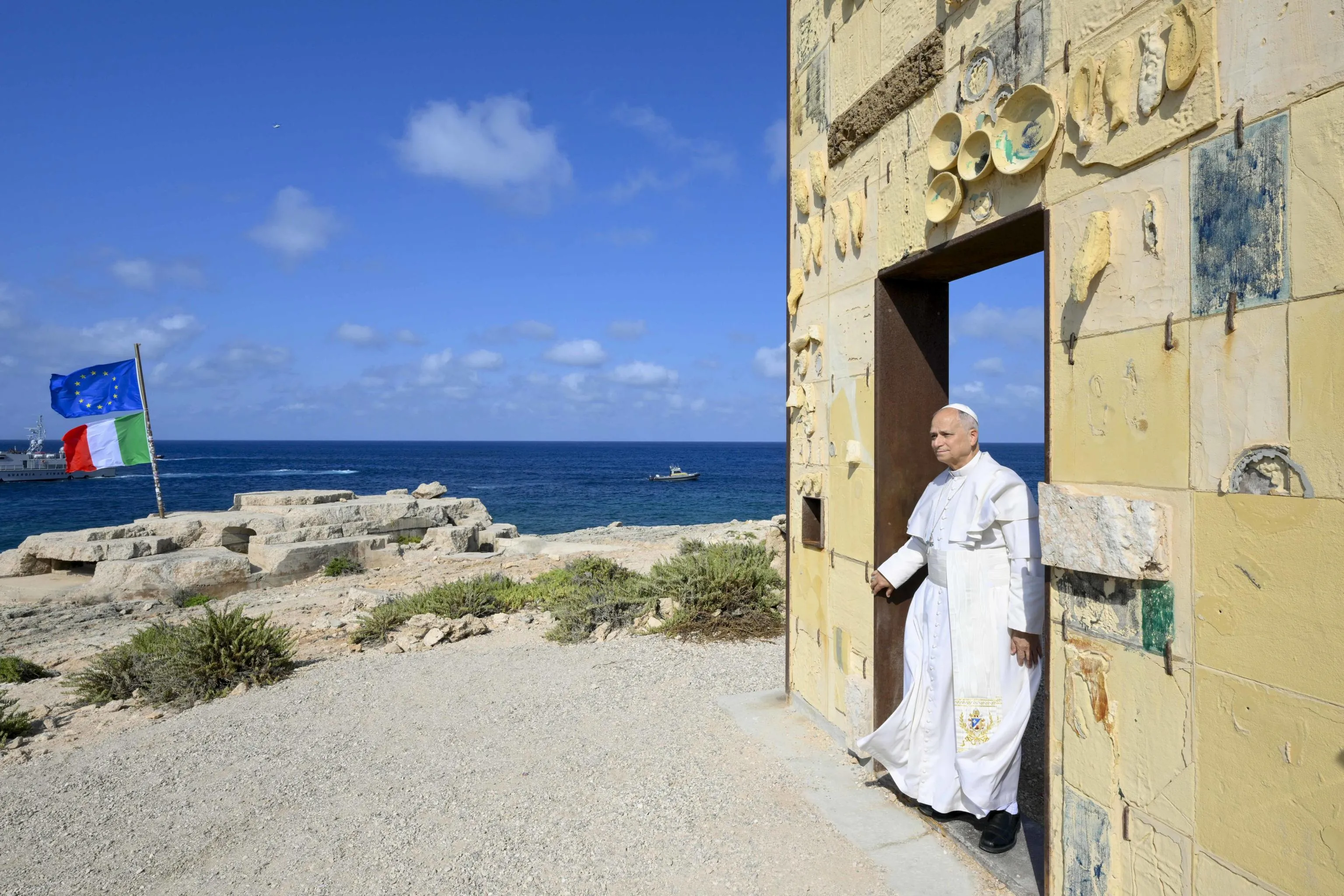 La visita di Papa Leone XIV a Lampedusa, il 4 luglio 2026. Prevost varca la Porta d’Europa, il monumento che rende omaggio ai migranti, simbolo della frontiera più a sud dell’Italia e del continente. Davanti a lui le bandiere italiana ed europea e quel mare teatro di continue tragedie / Ansa - Vatican media press office