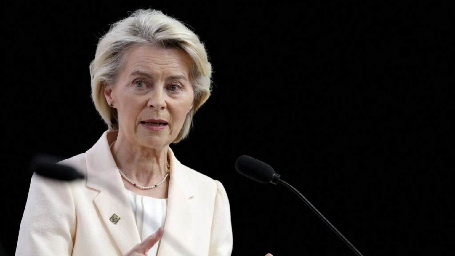 Una donna bionda, di mezza età, indossa un tailleur color crema e parla da un podio in legno. È ripresa a mezzobusto, con le mani sollevate mentre gesticola. Davanti a lei ci sono due microfoni neri; lo sfondo è completamente scuro, mettendo in risalto il