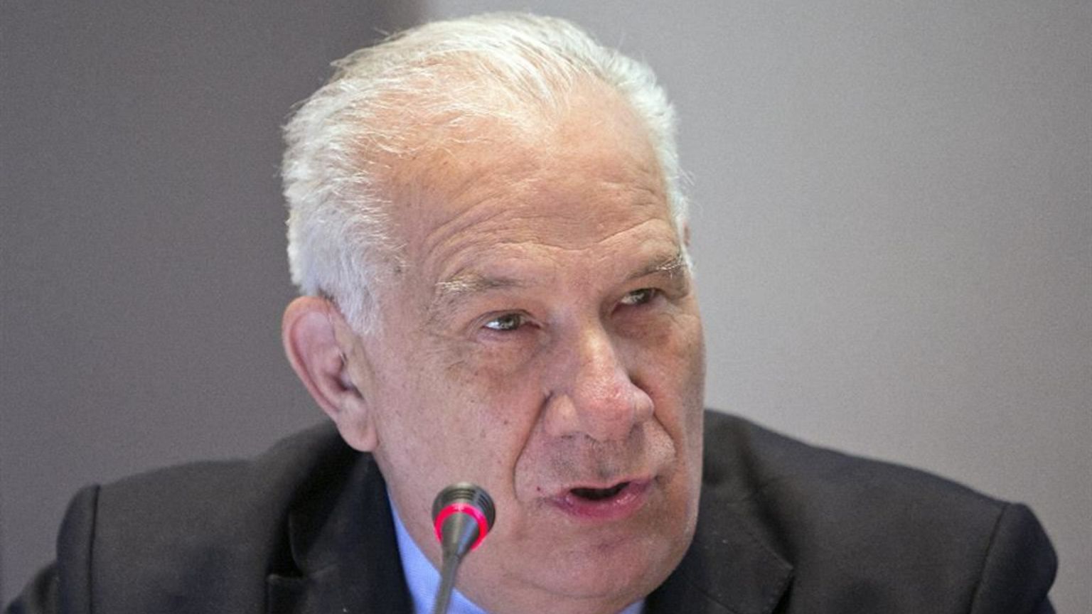 Primo piano di un uomo anziano dai capelli bianchi, in giacca e cravatta scura. È seduto a un tavolo e parla davanti a un microfono con una piccola luce rossa accesa. Lo sfondo è neutro; il suo volto è leggermente di profilo e l’espressione è concentrata.