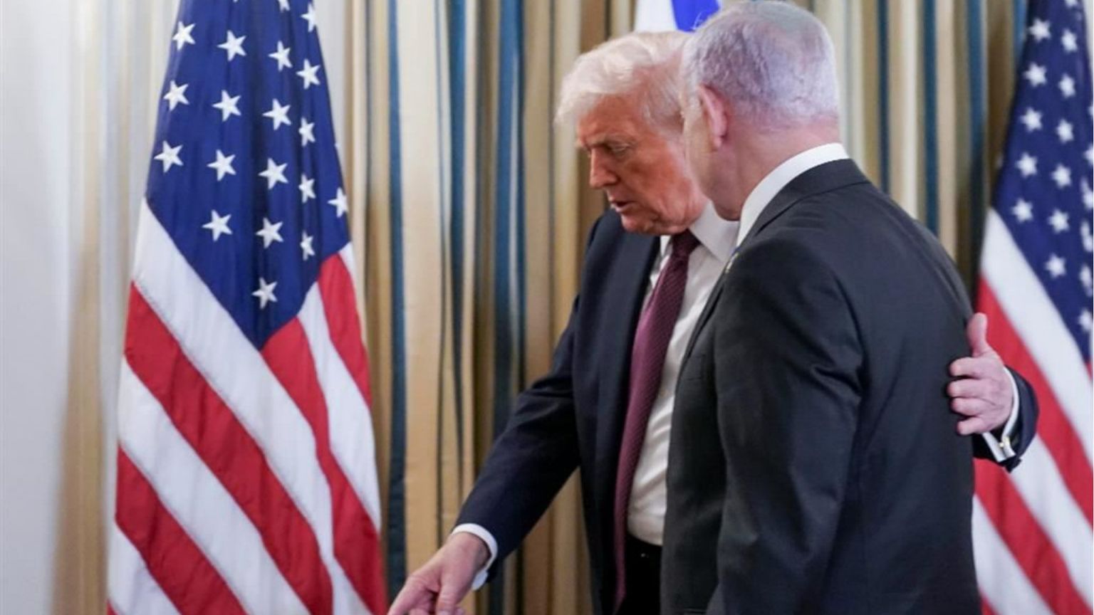 Due uomini in giacca e cravatta, di profilo, parlano da vicino in una stanza con tende a righe. Quello a sinistra, con capelli chiari, tiene una mano sulla spalla dell’altro e indica verso il pavimento; ai lati si vedono due bandiere degli Stati Uniti e,