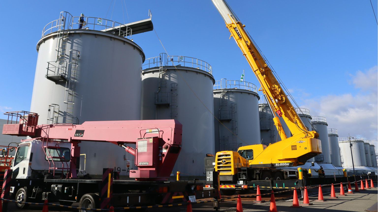 Il lungo braccio d'acciaio di una gru gialla si protende davanti alla prima di una serie di enormi cisterne cilindriche grigie, sotto il cielo azzurro